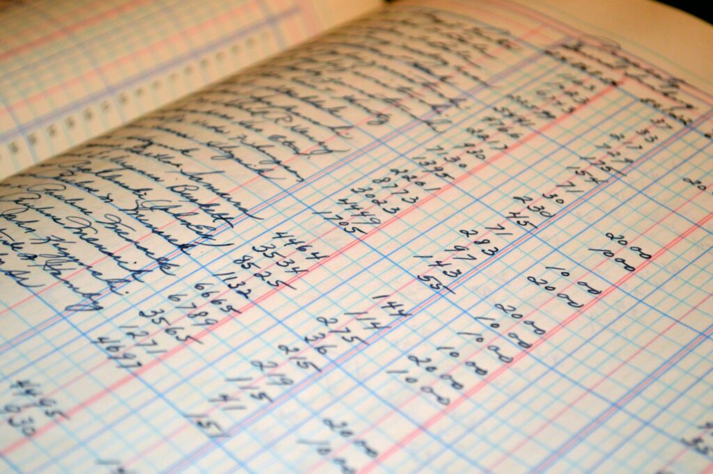 Close-up of a notebook page with blue grid lines and dense handwritten numbers and text in cursive, laid at an angle to the viewer.